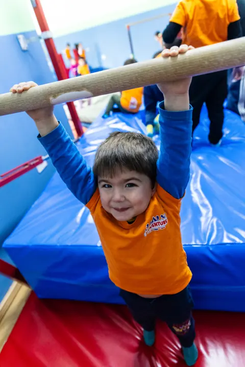 A smiling boy hanging from a bar over the mats
