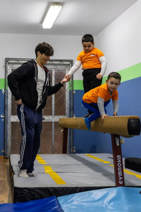 A coach helping two children along a balance beam
