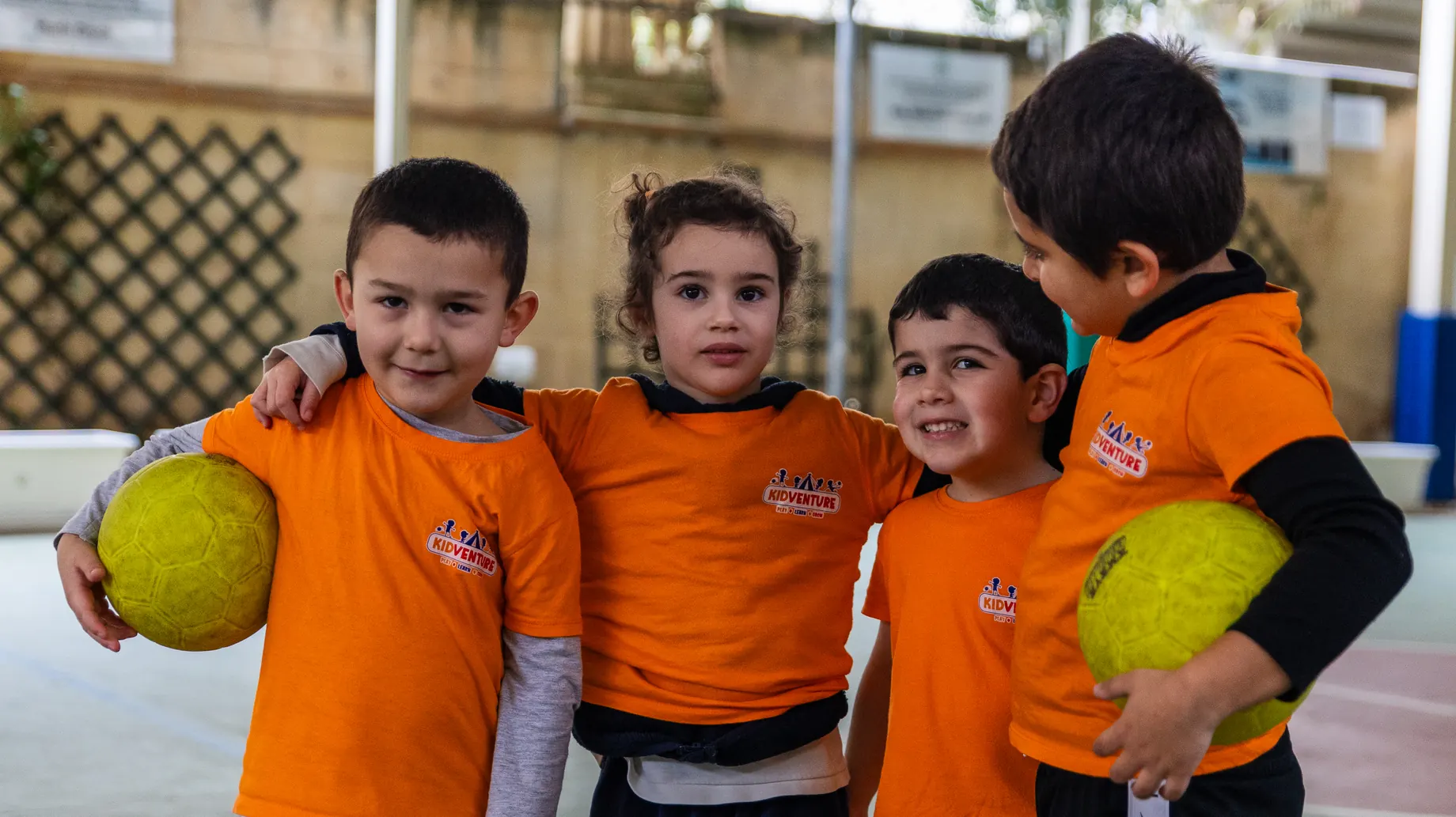 Four smiling friends in orange KidVenture T-shirts, arms around each other, two holding balls