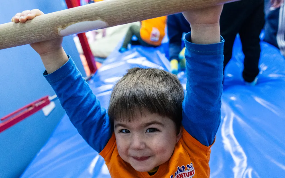 A smiling young boy in a KidVenture T-shirt hanging from a gymnastics bar